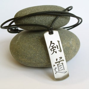 May include: A silver pendant necklace with a black leather cord. The pendant is rectangular and features the Chinese characters "剣道" (kendo) which means "the way of the sword".