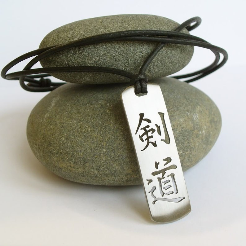 May include: A silver pendant necklace with a black leather cord. The pendant is rectangular and features the Chinese characters "剣道" (kendo) which means "the way of the sword".