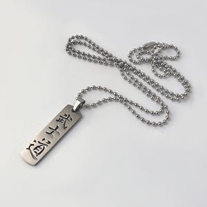 May include: A silver-toned rectangular pendant necklace featuring a ball chain and a metal tag engraved with the Japanese kanji characters for Bushido. The pendant has a smooth, metallic finish and is suspended from a matching silver-colored ball chain.
