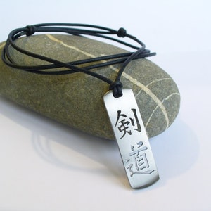 May include: A silver pendant necklace with a black leather cord. The pendant is rectangular and features the Japanese characters "剣道" (kendo) which means "the way of the sword".