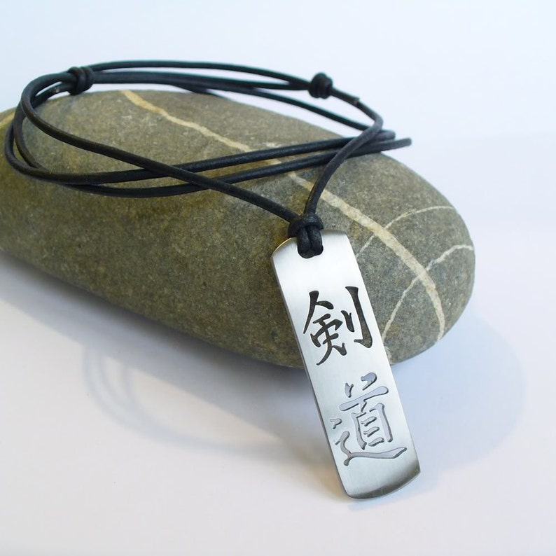 May include: A silver pendant necklace with a black leather cord. The pendant is rectangular and features the Japanese characters "剣道" (kendo) which means "the way of the sword".