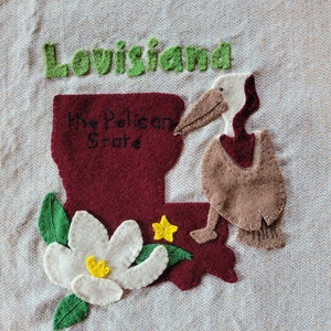May include: A felt applique depicting the state of Louisiana in burgundy, with the word "Louisiana" in green. A pelican, a magnolia flower, and a yellow star are also included. The text "The Pelican State" is written on the burgundy state shape.