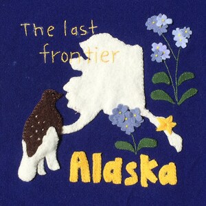 Pode incluir: Têxtil azul com um design de aplicação de feltro. O design inclui um contorno branco do Alasca, um pássaro marrom e branco, flores azuis e a palavra "Alaska" em amarelo. O texto "The last frontier" é bordado em amarelo.