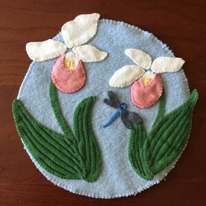 Könnte beinhalten: Ein runder, hellblauer Filz-Tischset mit zwei rosa-weißen Frauenschuhblüten, grünen Blättern und einer blau-grauen Libelle. Die Blüten haben gelbe Zentren und sind mit weißem Faden genäht.