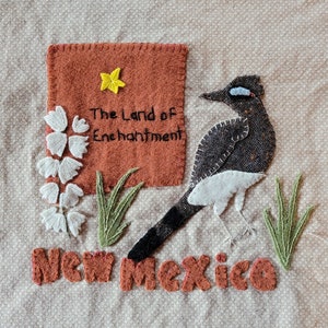 May include: A brown felt applique with the text "The Land of Enchantment" and "New Mexico" in white stitching. A white and gray bird applique with a yellow star is above the text.