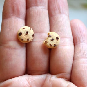 May include: Two miniature chocolate chip cookies, one whole and one with a bite taken out, rest on a person's palm. The cookies are small, round, and have a light brown color with dark brown chocolate chips.