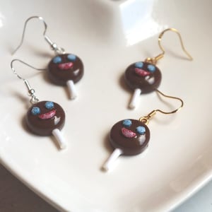 May include: Four pairs of novelty earrings shaped like chocolate lollipops with smiling faces. The earrings have brown, blue, pink, and white details. Two pairs have silver hooks, and two pairs have gold hooks. The earrings are on a white, heart-shaped plate.
