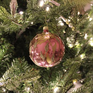 May include: A pink Christmas ornament with a gold floral design hangs on a green Christmas tree.