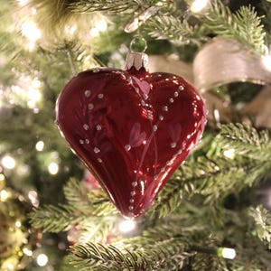 May include: A heart-shaped Christmas ornament in a deep red hue, adorned with small white dots and delicate floral designs. The ornament hangs from a Christmas tree with green needles and twinkling lights, creating a festive holiday scene.