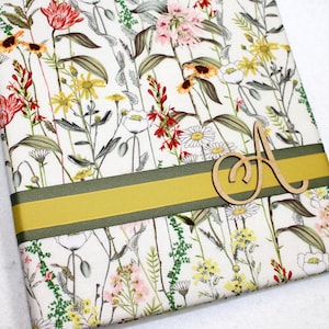 May include: A white fabric with a floral pattern in shades of red, yellow, and green. A yellow and green ribbon is wrapped around the fabric with a wooden letter "A" on top of the ribbon.