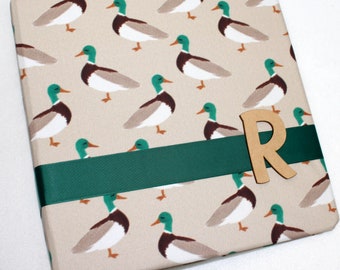May include: A beige gift box with a green ribbon and a wooden letter "R" on top. The box is covered in a pattern of brown and green ducks.