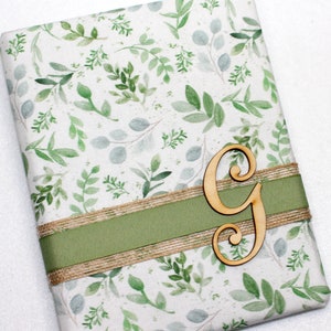 May include: A white gift wrap with a green leafy pattern and a gold ribbon. A wooden letter G is placed on top of the ribbon.
