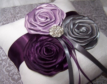 May include: A white satin ring pillow with three fabric roses in shades of purple, gray, and lavender. A rhinestone brooch is centered on the pillow.