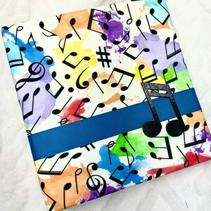 May include: A notebook with a musical theme. The cover features a white background with colorful watercolor splashes and black musical notes. A dark blue ribbon wraps around the notebook, and a black musical note cutout is attached.