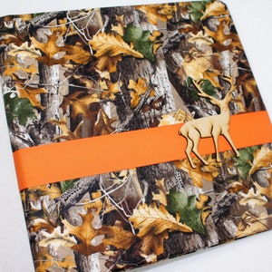 May include: A scrapbook with a camouflage pattern featuring brown, green, and tan leaves. The scrapbook has a bright orange ribbon around it and a wooden deer cutout on top of the ribbon.