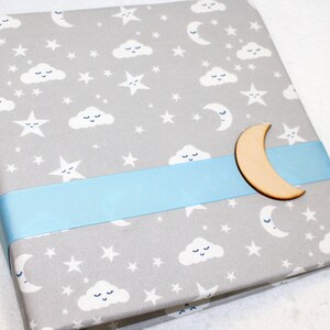 May include: A gray fabric photo album with a blue ribbon and a wooden crescent moon embellishment. The album features a repeating pattern of white clouds with faces and stars.