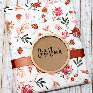 May include: A floral-patterned gift book with a white background and red, pink, and orange flowers. A wooden circle with the words "Gift Book" is centered on the book. A rust-colored ribbon wraps around the book.