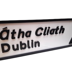 May include: A white sign with black lettering and a black border. The sign reads "Átha Cliath Dublin" with an arrow pointing right. The sign is angled slightly towards the viewer.