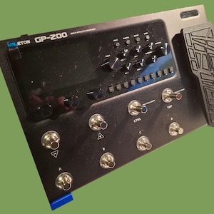 Puede incluir: Un procesador multiefectos Valeton GP-200 negro con pantalla integrada, numerosos botones y conmutadores de pie. Un pedal de expresión negro está adjunto. El dispositivo tiene detalles en azul y está diseñado para efectos de guitarra.
