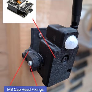 Op de afbeelding: Zwarte plastic camerabeugel met een witte sensor en antenne. De tekst "ESP32CAM niet inbegrepen" en "M3 Cilinderkopschroeven Inbegrepen" zijn zichtbaar. De beugel is bevestigd aan een houten oppervlak.