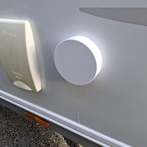 Op de afbeelding: Een witte ronde afdekplaat gemonteerd op een lichtgrijs voertuigpaneel, naast een beige Truma Ultraflow filterbehuizing. Een dunne verticale waterstreep loopt langs het paneel vanaf de onderkant van de ronde afdekplaat naar beneden.