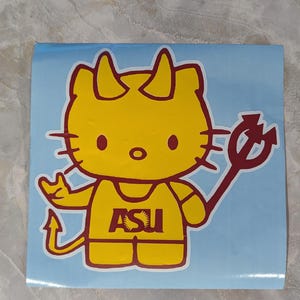 Puede incluir: Una pegatina amarilla con un gato de dibujos animados con cuernos de diablo y un tridente. El gato lleva un atuendo amarillo con las letras "ASU" en granate. El fondo es azul claro.