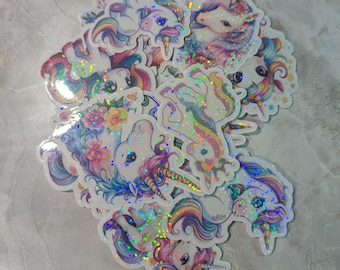 May include: A collection of holographic stickers featuring whimsical unicorn designs. The stickers showcase unicorns with rainbow manes and tails, adorned with flowers and glitter. The stickers have a shimmering, iridescent effect, adding a magical touch to the overall aesthetic.