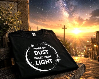 May include: Black t-shirt featuring a white circular design with the text "MADE OF DUST FILLED WITH LIGHT". The shirt is placed on a wooden surface, with a sunset, city, and cross in the background.