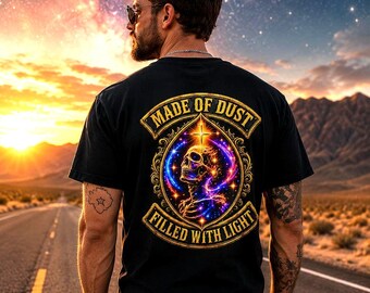 May include: Black t-shirt with a skull graphic and the text "MADE OF DUST FILLED WITH LIGHT". The design features a skull with a cross and colorful cosmic elements. The image is set against a sunset and road background.