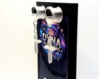 May include: Clear, cross-shaped earrings suspended from silver-coloured plugs. The earrings are displayed on a black stand with the text "DYNA 3D PRINTING" in a colourful, swirling design. The background is white.