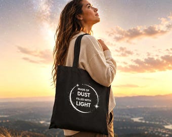 May include: Black tote bag featuring the text "MADE OF DUST FILLED WITH LIGHT" in a white circular design. The bag has a long handle. The image is set against a scenic background of a sunset and a starry sky.