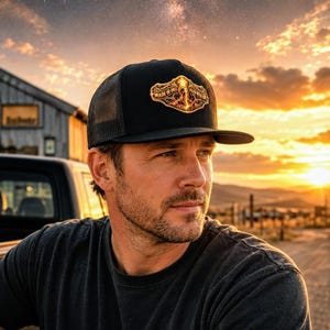 May include: A black trucker hat with a mesh back and a flat brim. The front panel features an ornate, glowing gold-toned patch with the text "MADE OF DUST" and "FALLEN WITH FIRE" displayed in a decorative, stylized font across the center.