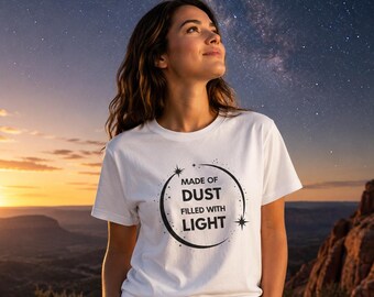 May include: White t-shirt featuring the text "MADE OF DUST FILLED WITH LIGHT" in a black circular design. The person is standing outside with a night sky backdrop.