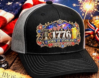May include: Black and grey trucker cap with a patriotic design. The cap displays the year "1776", the text "Born of Starlight, Forged by Sacrifice", and an illustration of the Liberty Bell and American flag. The background includes an American flag and a starry sky.