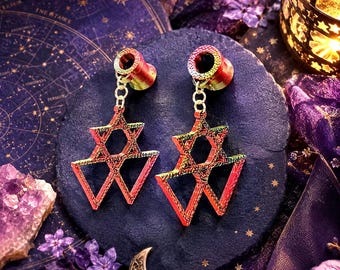 May include: A pair of iridescent red and gold star-shaped earrings. The earrings feature a Star of David design, connected to the ear tunnels by a delicate chain. The earrings are displayed on a dark blue surface with celestial-themed decorations.
