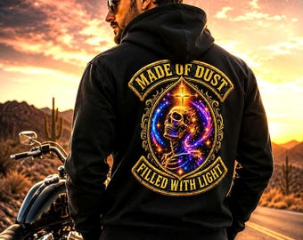 May include: Black hoodie with a skull and cross graphic, and the words "MADE OF DUST" and "FILLED WITH LIGHT" in gold. The person is wearing sunglasses and standing near a motorcycle on a desert road at sunset.