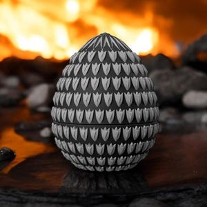 May include: A black and white decorative egg-shaped object with a detailed pattern of stylized leaf shapes. The object is set against a blurred background of rocks and fire, creating a dramatic contrast.