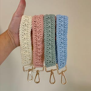 May include: Four crochet bag straps in cream, pink, sage green, and light blue. Each strap features a starburst pattern and gold-colored metal clasps. The straps are held up against a neutral background.