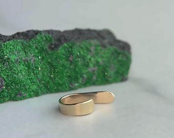 Könnte beinhalten: Ein polierter goldfarbener Metallring mit einem offenen Bypass-Band und einer glatten, reflektierenden Oberfläche. Der Ring besteht aus einer einfachen, durchgehenden Metallschleife mit abgerundeten Enden und zeigt ein schlichtes Design ohne Steine.
