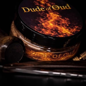 May include: A jar of Dude of Oud small batch artisan shave soap with a black lid and a label featuring a fiery design. The jar is filled with a brown substance, and the text "Dude of Oud" is in gold.