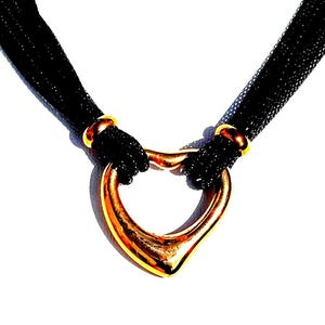 May include: A necklace featuring multiple thin black cords gathered by two gold-toned metal beads. The cords pass through a central, smooth, gold-toned heart-shaped pendant. The design is symmetrical, with the black cords extending upwards.