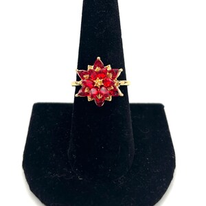 May include: A gold-toned ring featuring a cluster of red gemstones arranged in a floral pattern. The ring is displayed on a black velvet ring holder against a white background. The gemstones are a deep red color.