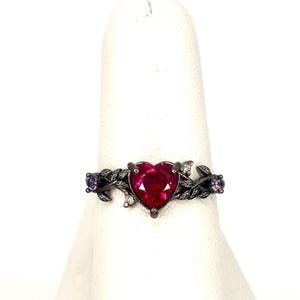 May include: A black metal ring featuring a central heart-shaped red gemstone. The band is designed with a textured leaf motif and accented with small clear stones and two round purple gemstones on the sides, creating a detailed botanical look.
