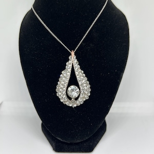 May include: A silver teardrop-shaped pendant necklace with a large clear crystal in the center. The pendant is surrounded by smaller clear crystals.