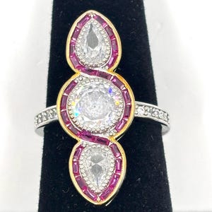 May include: A silver ring with a gold-toned setting, featuring three clear, sparkling stones. The central stone is round, while the top and bottom stones are pear-shaped. The ring is accented with small, red stones and additional clear stones on the band.