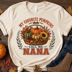 May include: A cream-colored short-sleeve t-shirt featuring a graphic of a woven basket filled with pumpkins, a sunflower, wheat, and autumn leaves. The text reads "MY FAVORITE PUMPKINS CALL ME NANA" in orange and green lettering with a leaf border.