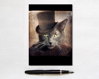 Può includere: Biglietto di auguri con un'immagine color seppia di un gatto grigio con un cappello a cilindro marrone. Il gatto ha occhi verdi e baffi bianchi. Il biglietto ha un bordo nero. Una penna nera è in primo piano.