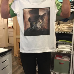 May include: White t-shirt with a vintage-style image of a cat wearing a top hat. The cat has gray fur and yellow eyes. The image is printed on the front of the shirt.