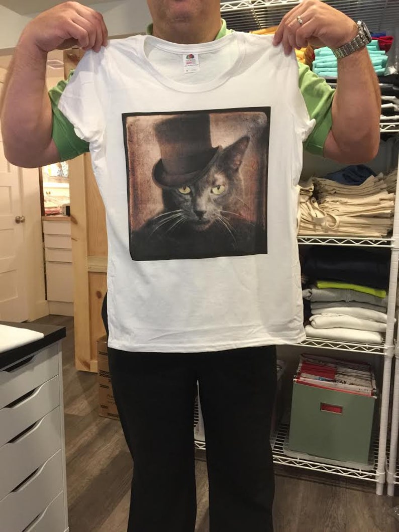 May include: White t-shirt with a vintage-style image of a cat wearing a top hat. The cat has gray fur and yellow eyes. The image is printed on the front of the shirt.