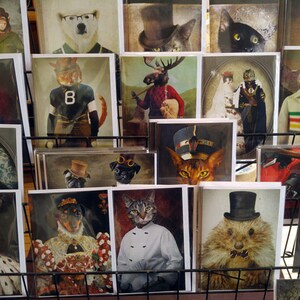 Puede incluir: Una colecci&oacute;n de impresiones de arte enmarcadas con varios animales vestidos con ropa. Las impresiones se muestran en un estante de metal negro. Algunos de los animales incluyen una ardilla, un gato, un perro, un alce y un tej&oacute;n.